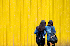 Two dark-haired girls wearing backpacks face away from the camera, looking at a yellow wall.