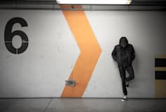 
        A hooded man leans against a wall, his hat tipped down to block his face.
        He's in an industrial setting. The wall behind him has a large number 6
        painted on it, as well as an orange, zig-zag, caret shape.
      