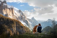 A woman and a golden retriever in the mountains. They're sitting, facing away from the camera while the dog noses the woman. They're surrounded by natural splendor including majestic mountains, puffy clouds, and evergreen-studded hills.