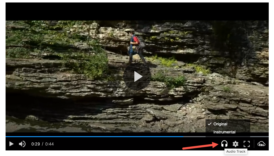 Cloudinary Video Player with a red arrow pointing at the Audio Track icon, which show a popup with a checkmark next to Original. The other option is Instrumental. The video still shows someone hiking up a mountain.