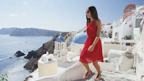 A woman in a red dress climbing stairs in a beautiful, ancient, white-walled Greek village by the sea.